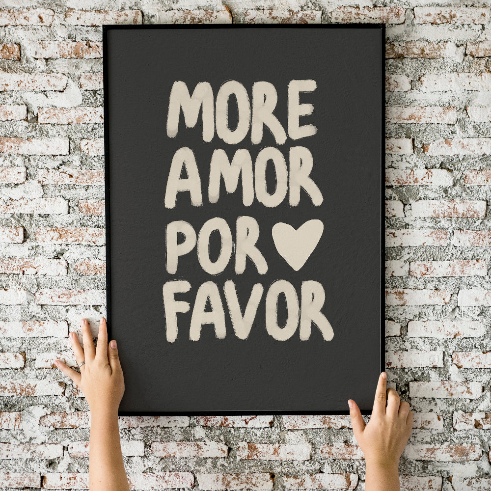 Person hanging a framed poster with 'More Amor Por Favor' text on a brick wall.